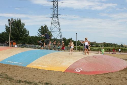 Children playing on a large, colorful inflatable bounce pad at a park, with trees and power lines in the background.