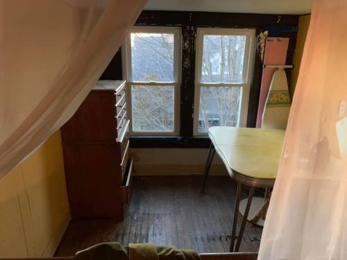 A cozy room with two windows, a yellow table, a dresser, and an ironing board, partially obscured by a curtain.