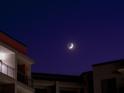 A crescent moon shines in a deep blue sky above modern apartment buildings at dusk.