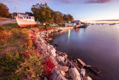 A serene waterfront scene at sunset, featuring a boat on land, rocky shore, and calm water with distant boats.