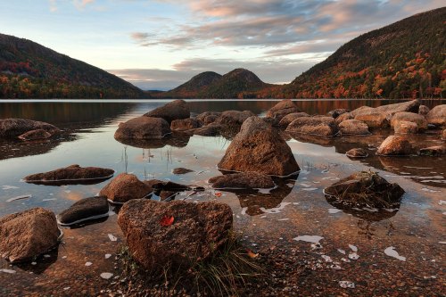 A serene lake surrounded by mountains, with large rocks in the foreground and autumn foliage reflecting in the water.