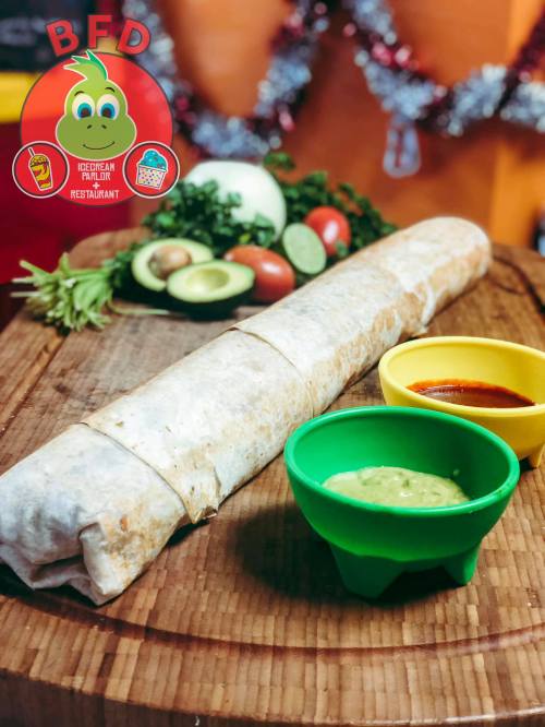 A large burrito on a wooden board, accompanied by green and yellow bowls of salsa and guacamole, with fresh ingredients in the background.