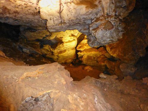 A dimly lit cave interior with rocky walls and a sandy floor, showcasing natural formations and shadows.