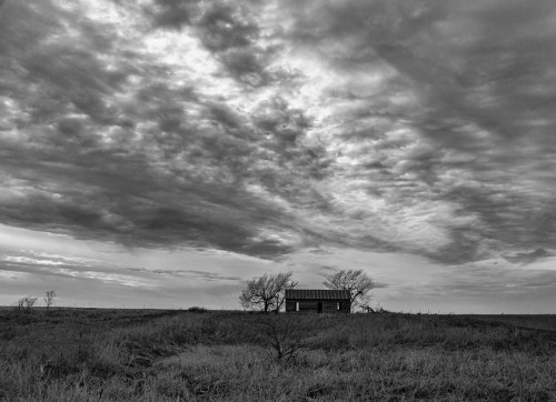 A solitary building stands in a grassy field under a dramatic, cloudy sky in black and white.