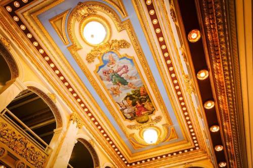 Ornate ceiling with intricate paintings and decorative moldings, featuring warm lighting and elegant architectural details.
