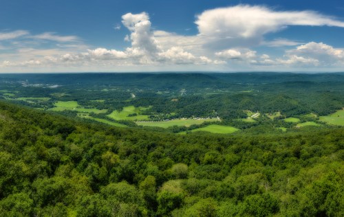 A panoramic view of lush green hills and valleys under a bright blue sky with scattered clouds.
