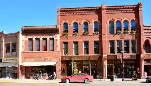 Historic brick buildings line a sunny street, featuring shops and decorative windows, with a red car parked in front.