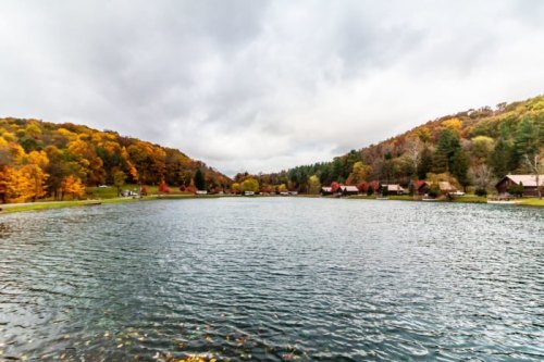 A serene lake surrounded by colorful autumn trees and quaint cabins under a cloudy sky.