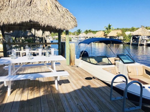 A wooden dock with a thatched roof, picnic table, and a boat by the water, surrounded by palm trees and clear blue skies.