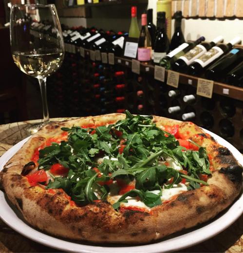 A delicious pizza topped with arugula and tomatoes, served with a glass of white wine in a cozy wine cellar setting.