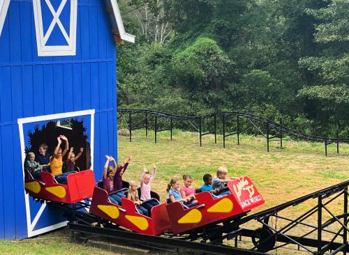 A colorful roller coaster exits a blue barn, with excited children raising their hands as they ride.