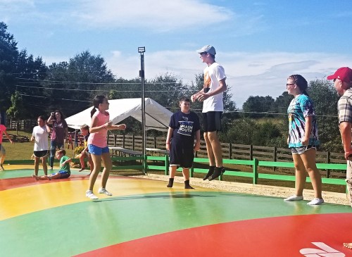 A group of children and teens play on a colorful inflatable bounce pad under a clear blue sky.