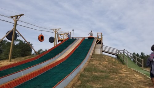 A large, green and orange slide on a hill, with a person preparing to go down and others nearby.