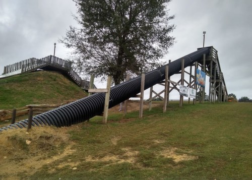 A large, black slide made of tubing descends from a wooden structure, surrounded by grass and trees under a cloudy sky.