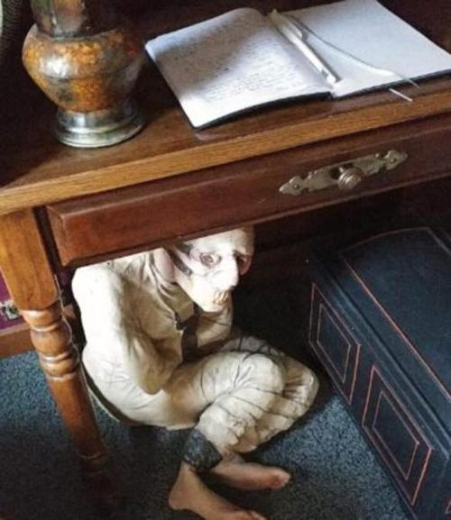 A creepy figure crouches under a desk, partially hidden, with a notebook and lamp nearby.