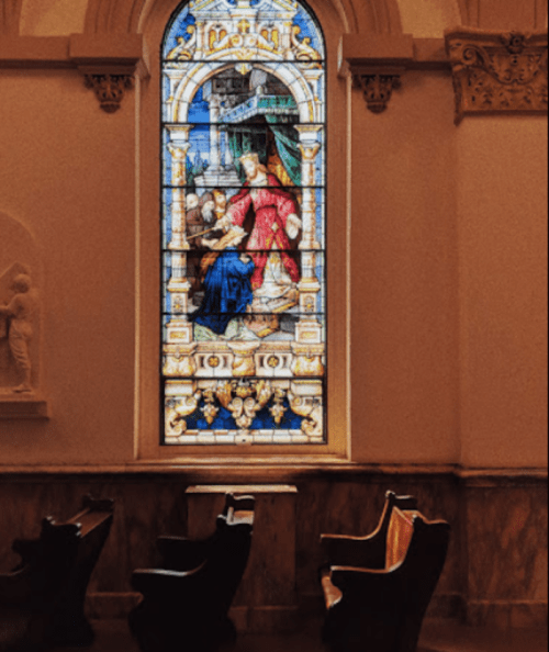 Stained glass window depicting a religious scene, framed by ornate architecture and wooden chairs below.