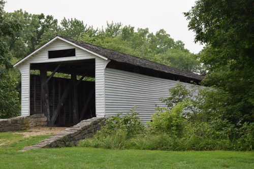 A white covered bridge stands beside a grassy area, surrounded by trees and a stone foundation.