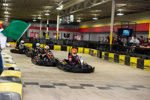 Go-kart racers compete on an indoor track, with one driver in the lead and a flag being waved in the background.