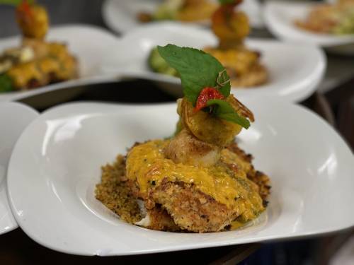A plated dish featuring a crispy seafood patty topped with shrimp, cheese, and garnished with herbs on a bed of grains.