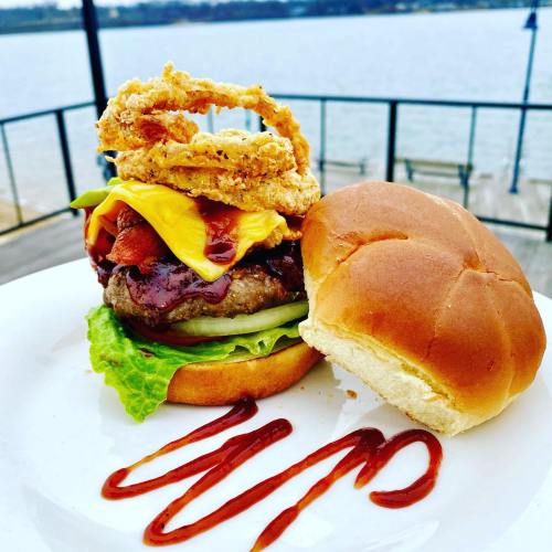 A delicious burger topped with cheese, bacon, lettuce, and crispy onion rings, served on a plate by the water.