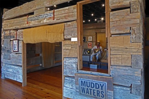 A wooden exhibit featuring a musician playing guitar, with a sign reading "Muddy Waters" and rustic decor.