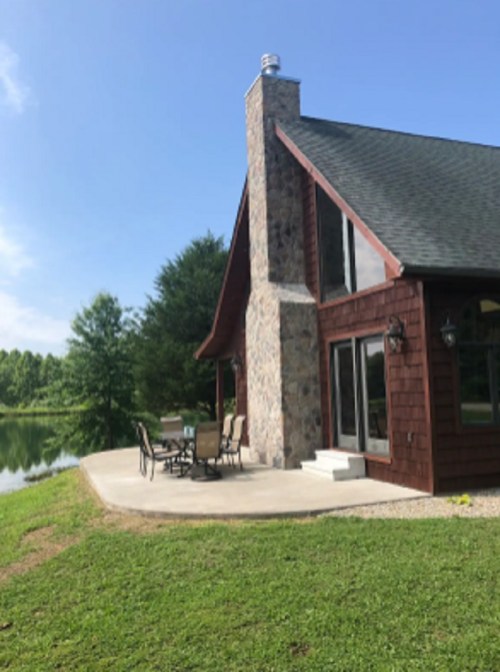 A rustic house with a stone chimney overlooks a serene pond, featuring a patio with outdoor furniture.