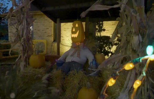 A scarecrow with a jack-o'-lantern head sits among hay bales and pumpkins, illuminated by festive lights.