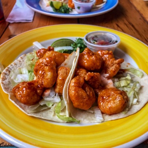 Two shrimp tacos topped with spicy sauce, lettuce, and a lime wedge, served with a side of salsa.