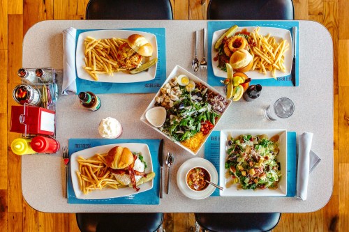 A table set with various dishes: burgers, fries, salads, and a dessert, all arranged neatly on colorful plates.