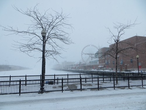 Snowy scene by a waterfront with bare trees, a ferris wheel in the background, and a foggy atmosphere.