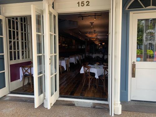 View of a restaurant entrance with open double doors, revealing a dimly lit dining area with tables and chandeliers.