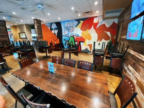 A restaurant interior with empty tables and chairs stacked upside down, featuring a colorful mural on the wall.