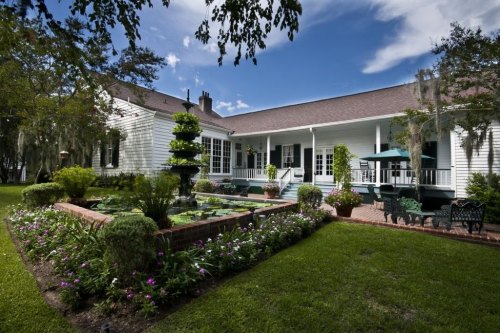 A charming white house with a garden, fountain, and patio seating under a blue sky with scattered clouds.