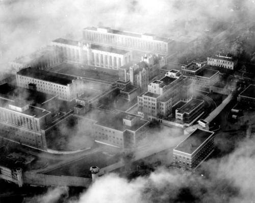 Aerial view of a large, industrial complex shrouded in fog, featuring multiple buildings and a surrounding wall.