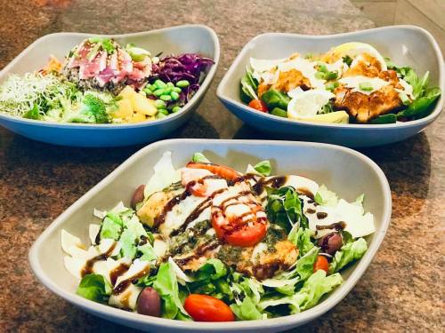 Three colorful salad bowls featuring greens, grilled chicken, tomatoes, and various toppings on a granite countertop.