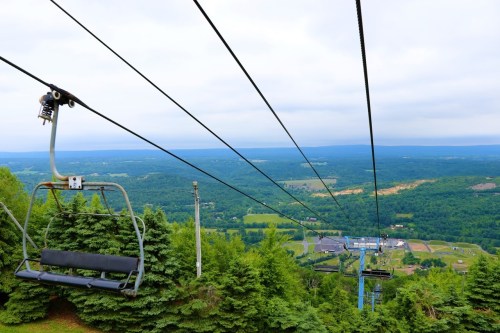 A chairlift ascends a mountain, offering a view of lush green valleys and distant hills under a cloudy sky.