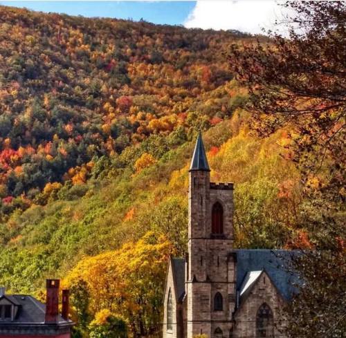 A stone church stands in front of a vibrant autumn landscape with colorful trees on a hillside.