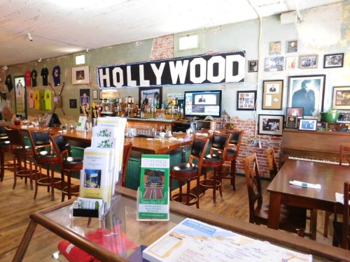 Interior of a bar with a "HOLLYWOOD" sign, wooden furniture, a piano, and various framed photos and memorabilia on the walls.