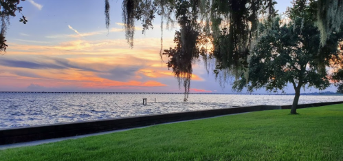 A serene waterfront scene at sunset, featuring a grassy area, trees, and a colorful sky reflecting on the water.