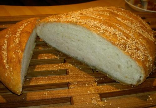 A loaf of freshly baked bread with a golden crust, sprinkled with oats, partially sliced on a wooden cutting board.