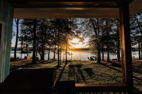Sunrise over a tranquil lake, framed by trees, casting long shadows on the ground. A peaceful morning scene.