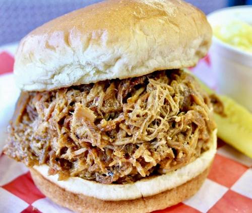 A close-up of a pulled pork sandwich on a bun, served with a side of coleslaw and a pickle.