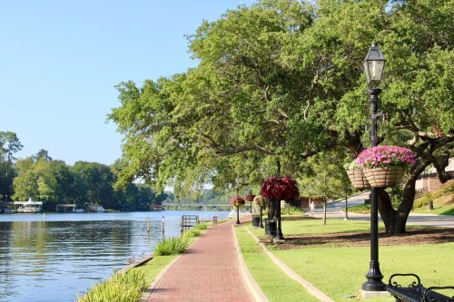 A scenic riverside path lined with trees and lampposts, featuring hanging flower baskets and calm water.