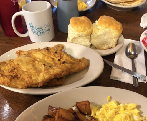 A plate of fried fish with biscuits, scrambled eggs, and potatoes, alongside a coffee cup on a wooden table.