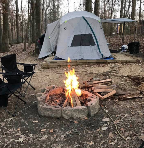 A campfire burns in a stone ring near a tent and camping chairs in a wooded area.