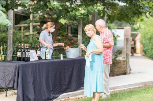 A woman in a blue dress holds a drink while a man in a red shirt stands nearby, as a server prepares drinks at a table.