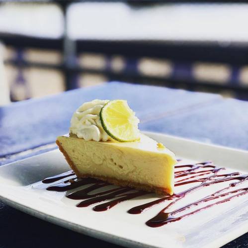 A slice of lime cheesecake topped with whipped cream and a lime slice, drizzled with chocolate sauce on a white plate.