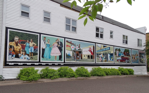 A colorful mural on a building featuring vintage scenes, cars, and people in retro clothing. Green bushes in the foreground.