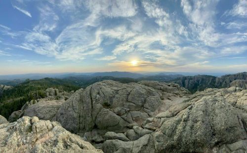 Panoramic view of rocky terrain under a colorful sky at sunset, with distant mountains and forests in the background.