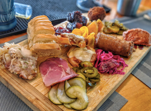 A wooden platter filled with various meats, pickles, bread, and colorful sides, showcasing a variety of textures and flavors.
